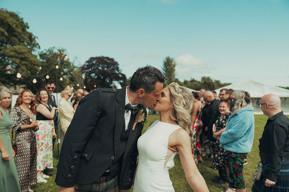Bride and groom share a kiss outdoors surrounded by wedding guests, the groom in tartan trews and the bride in a fitted sleeveless gown