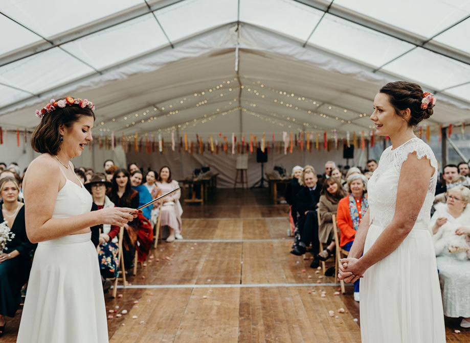 bride on left wearing a flower crown reads from a clipboard during a wedding ceremony as second bride on right looks on emotionally with hands clasped. Guests sitting on rows of wooden folding chairs look on. There are red, orange and pink streamers hanging from the marquee ceiling in the background