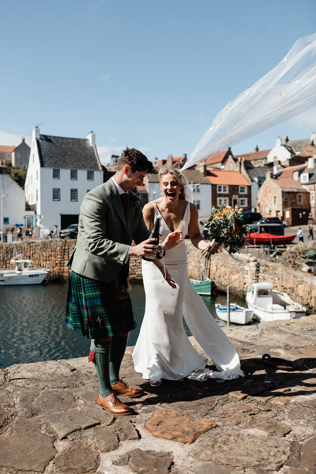 Bride and groom laughing as they pop a bottle of champagne by the harbour, her veil blowing in the wind