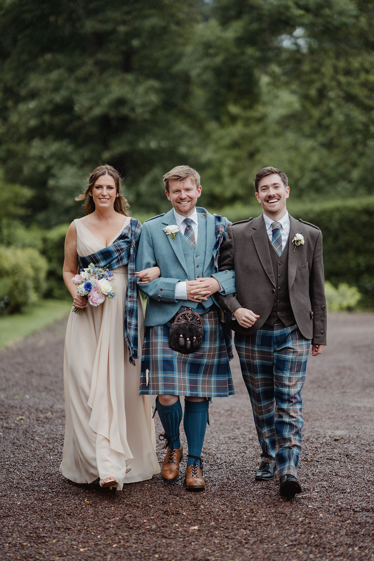 The other groom walks arm in arm with two wedding party members down a tree-lined path, all smiling and dressed in coordinated tartan outfits