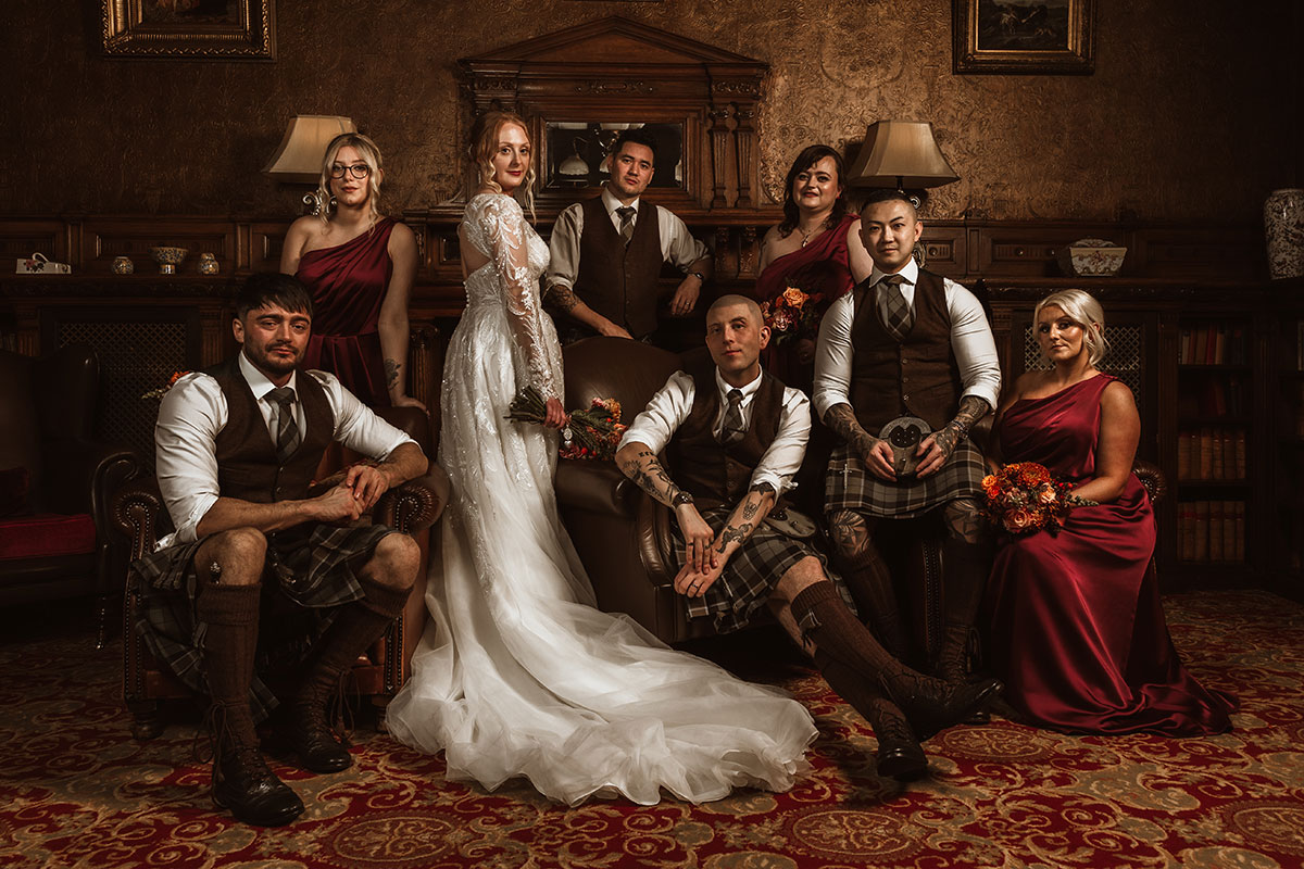 Bride and groom posing with their wedding party in an ornate room with vintage decor, groomsmen in kilts and bridesmaids in burgundy dresses