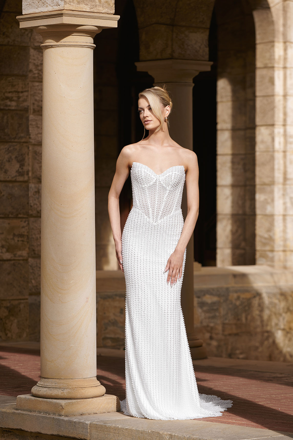 Bride in a strapless pearl-embellished column wedding dress with fitted silhouette and subtle sweetheart neckline, posed beside a stone column in an architectural setting