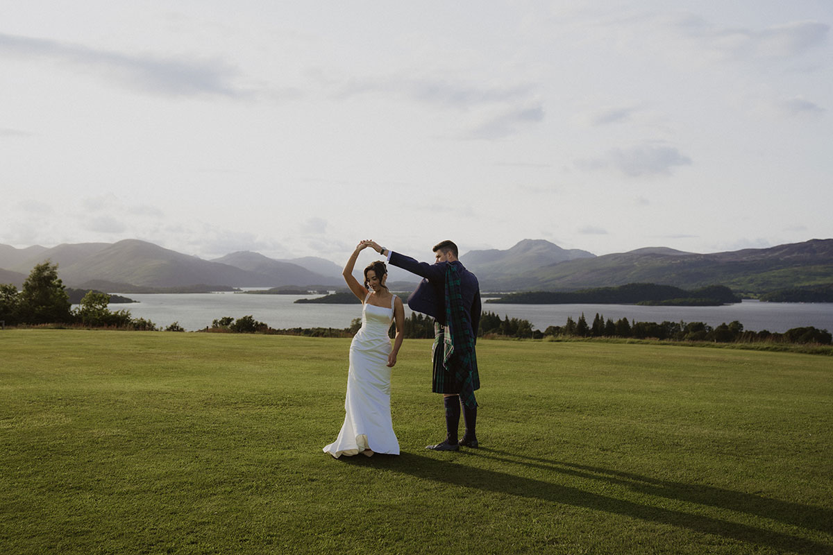 Bride and groom twirling on lawn overlooking Scottish loch and mountains during golden hour wedding portraits