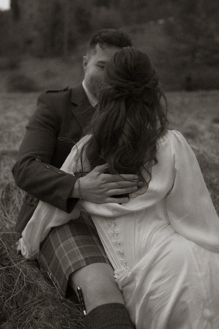 black and white photo of dark haired bride in a corset dress, with her back to the camera, facing groom in kilt outfit who has a hand softly in the middle of her back