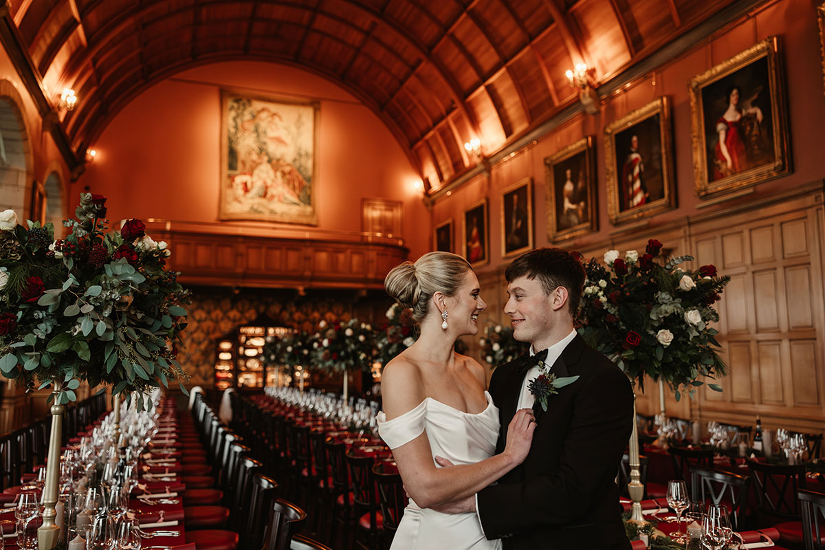 Couple standing together in Barnbougle Castle’s Minstrels’ Hall, surrounded by tall winter floral arrangements and long banquet tables.