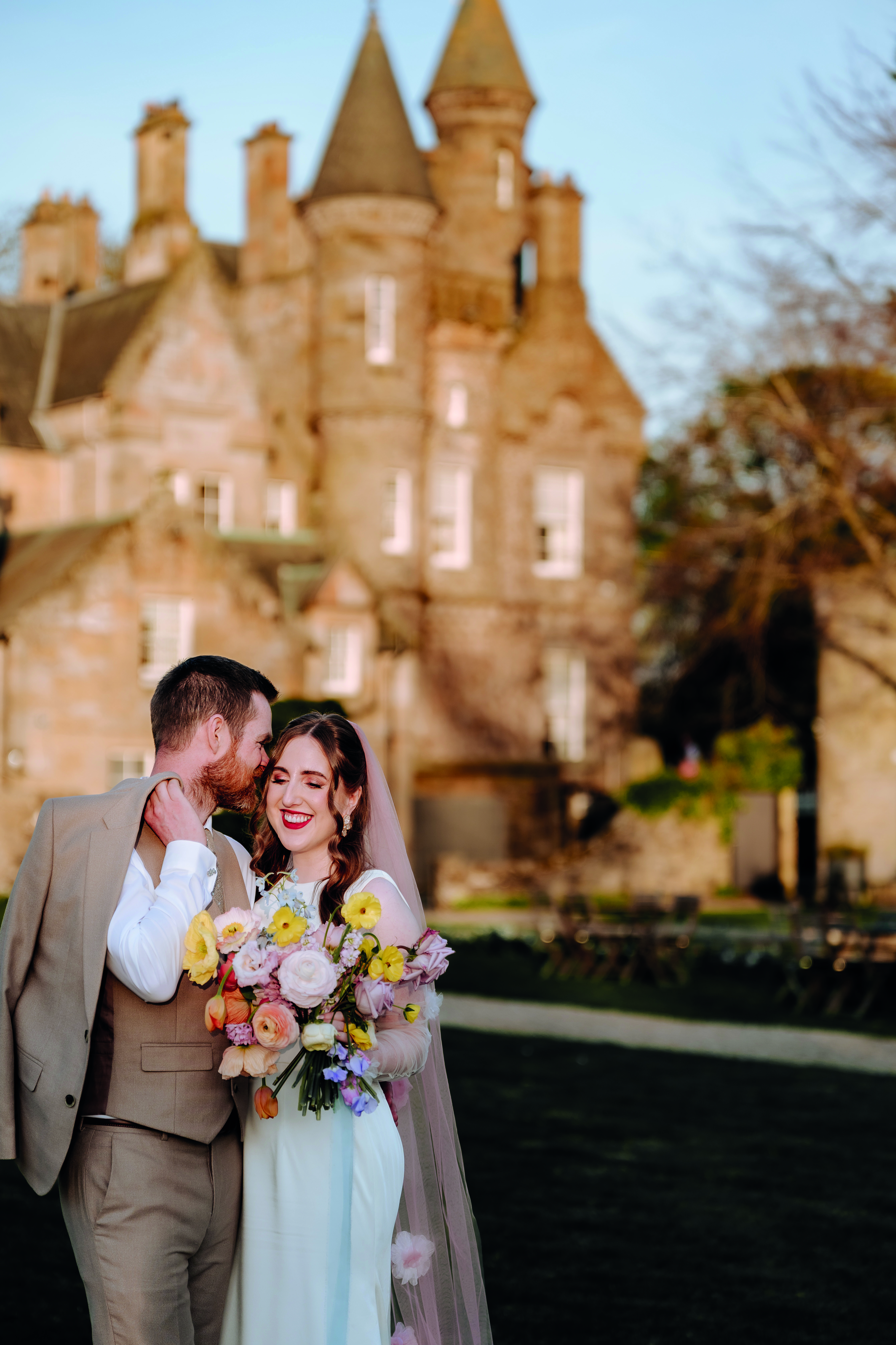 bride and groom smile outside carlowrie castle while bride wears pink floral wedding veil