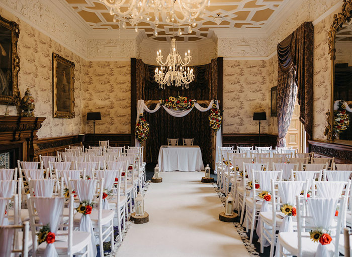 A room set up for a wedding ceremony with rows of white wooden chairs and a wooden arch at the end of the aisle with chiffon and flowers draped on it