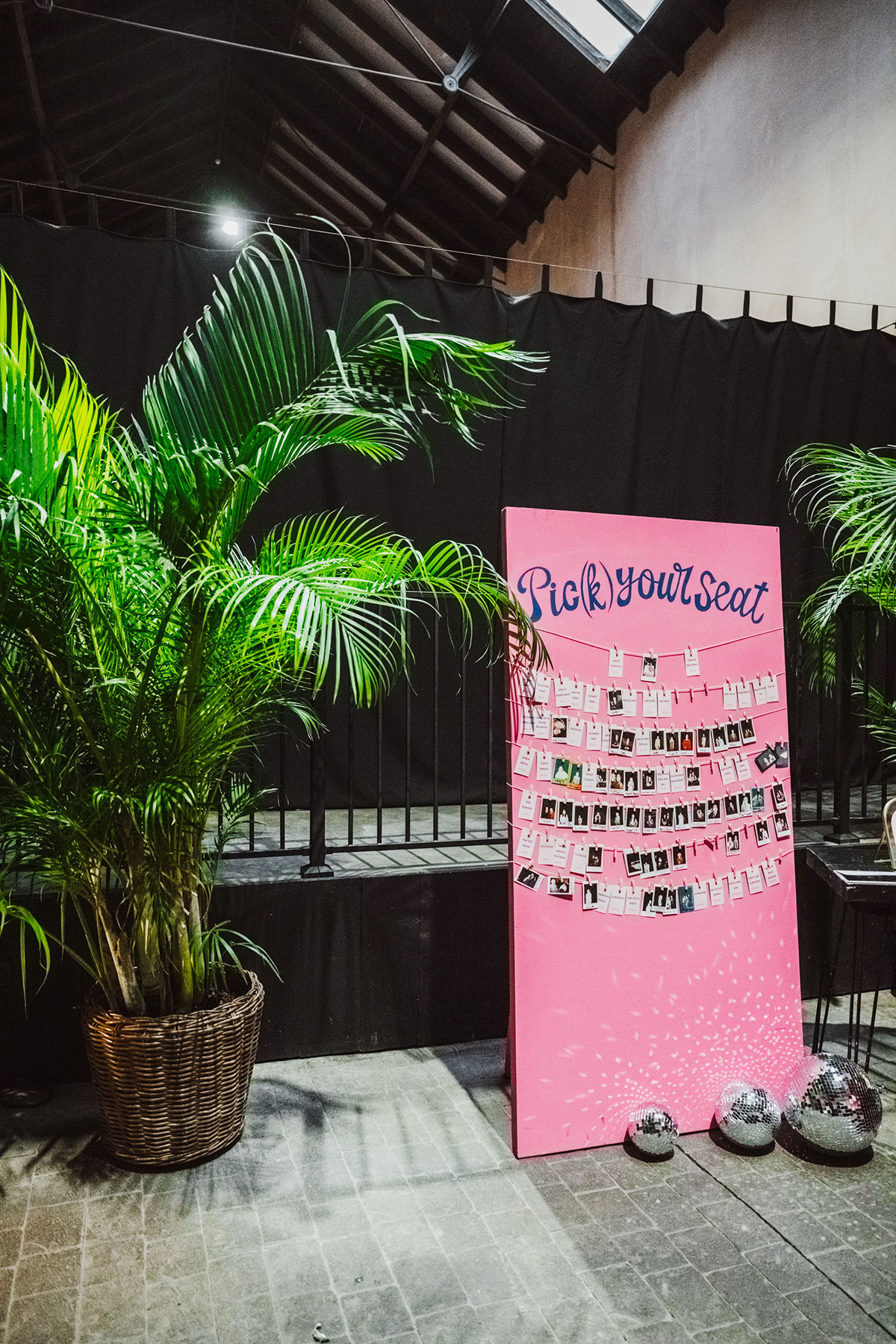 Bright pink “Pick Your Seat” wedding seating chart board decorated with rows of clipped Polaroid-style photos, placed beside tall potted palms and scattered disco balls.