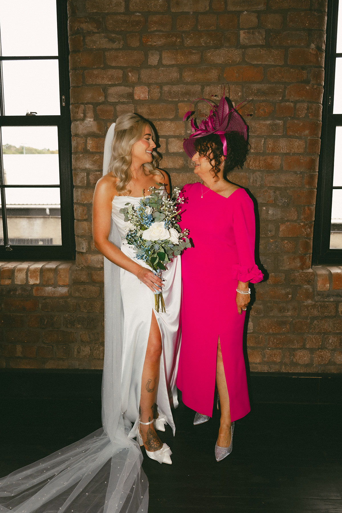 Bride in a white gown holding a bouquet stands beside an older woman in a bright pink dress and fascinator in front of a brick wall, both smiling at each other