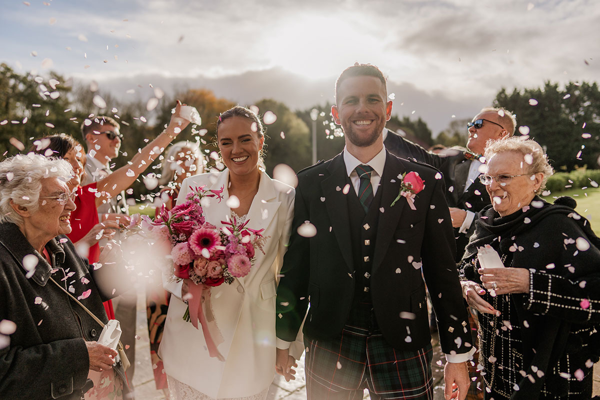 Bride in white suit and groom in tartan kilt walk through confetti after outdoor Scottish wedding ceremony