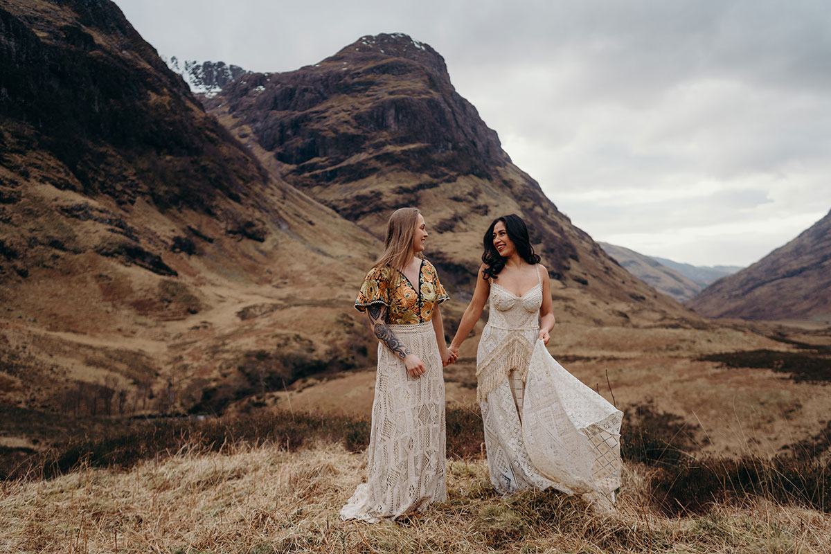 Couple walking hand in hand during a Scottish elopement shoot in Glencoe, wearing boho lace wedding outfits against a mountain backdrop.