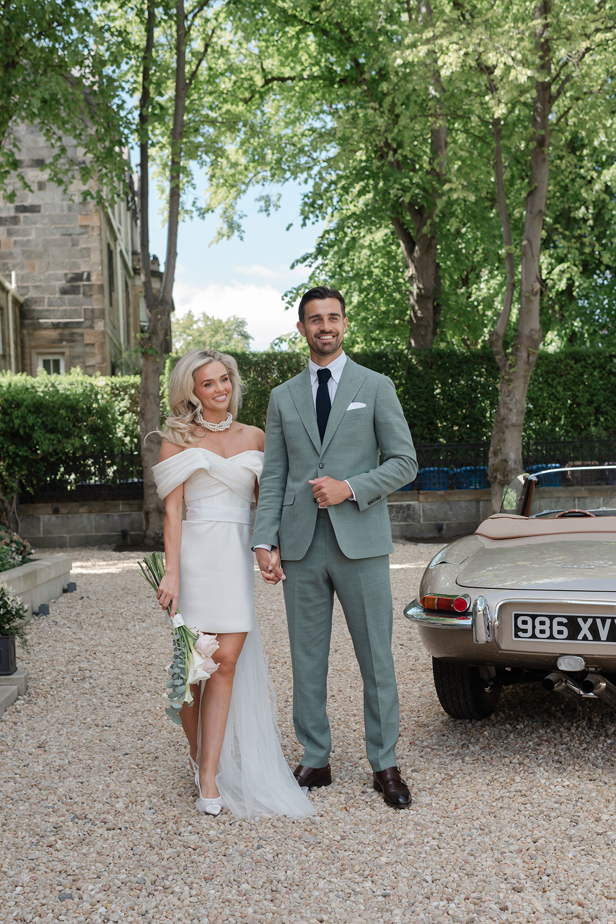 Bride in short off-the-shoulder wedding dress with tulle train and groom in sage green suit pose beside vintage car
