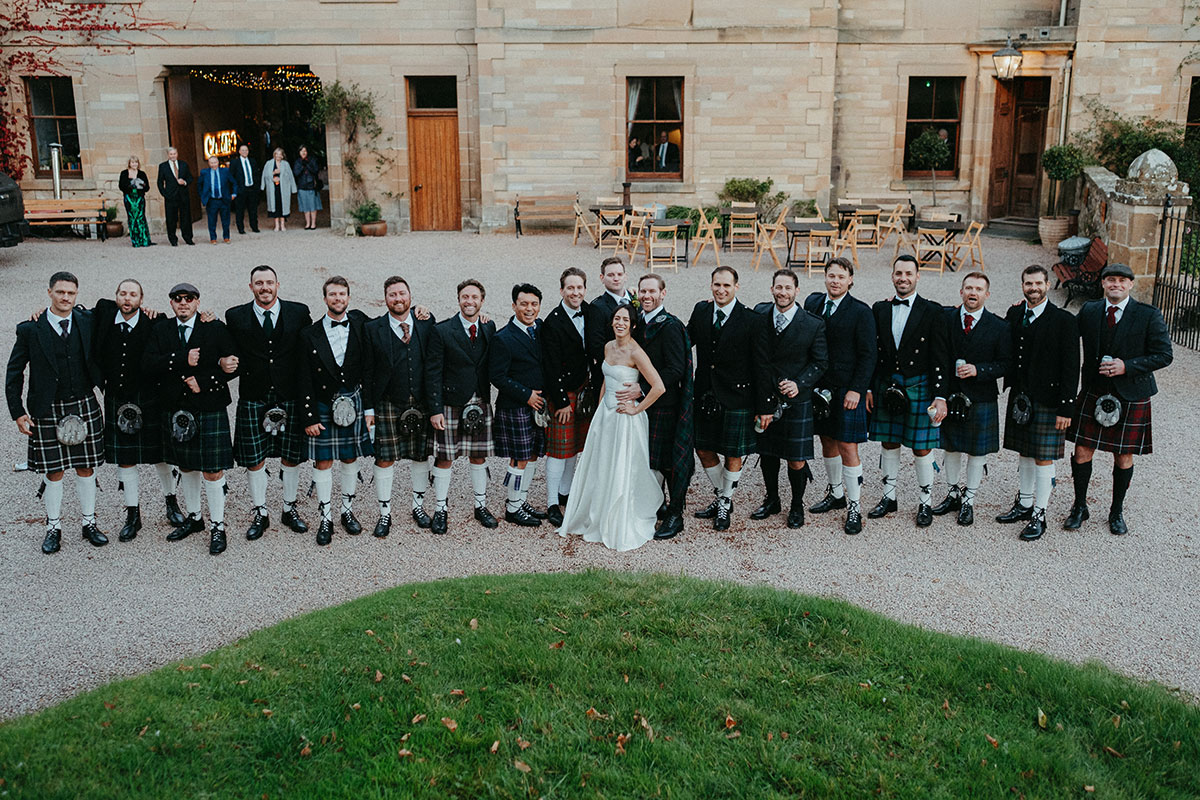 Bride and groom pose with wedding guests wearing traditional Scottish kilts in the courtyard of Cambo Country House.