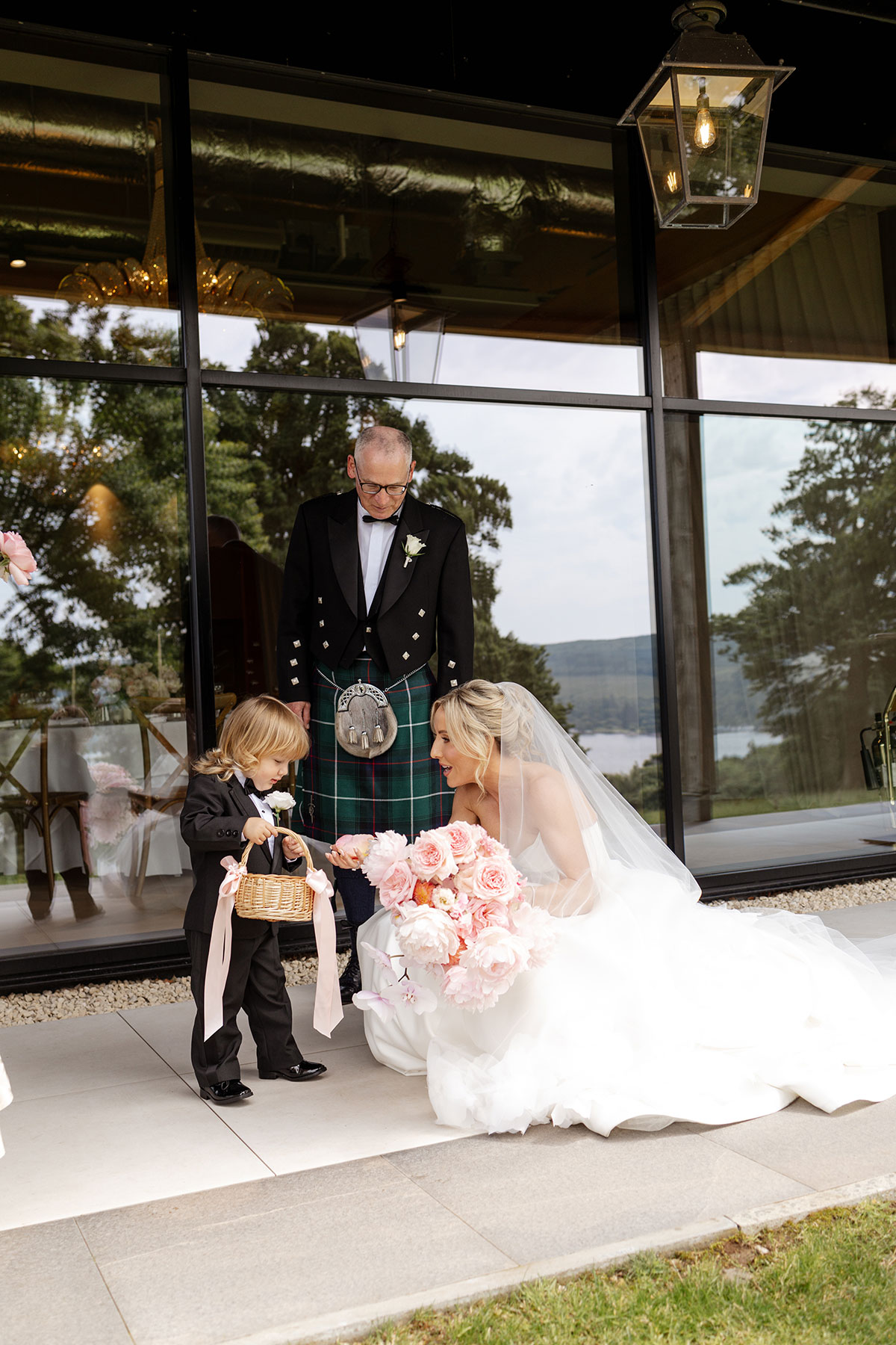 Bride crouches beside her young son holding a confetti basket outside Boturich Castle before the Loch Lomond wedding ceremony.