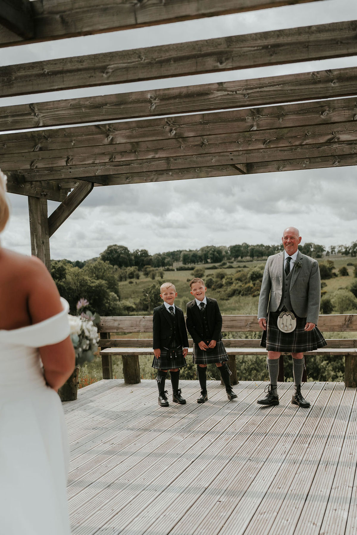 Groom and two boys in kilts smile as the bride approaches them on a wooden deck.