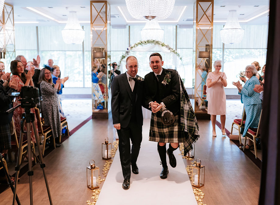 two elated-looking grooms holding hands walking up a white carpet aisle as rows of wedding guests either side clap and cheer