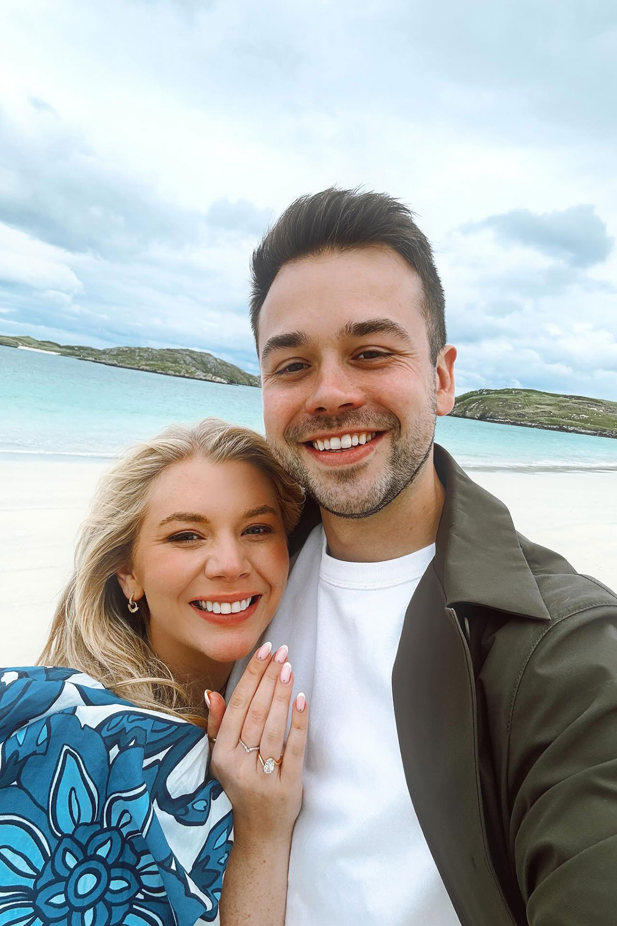 Smiling engaged couple taking a selfie on a white-sand Scottish beach, bride showing diamond engagement ring with turquoise sea and cloudy sky behind