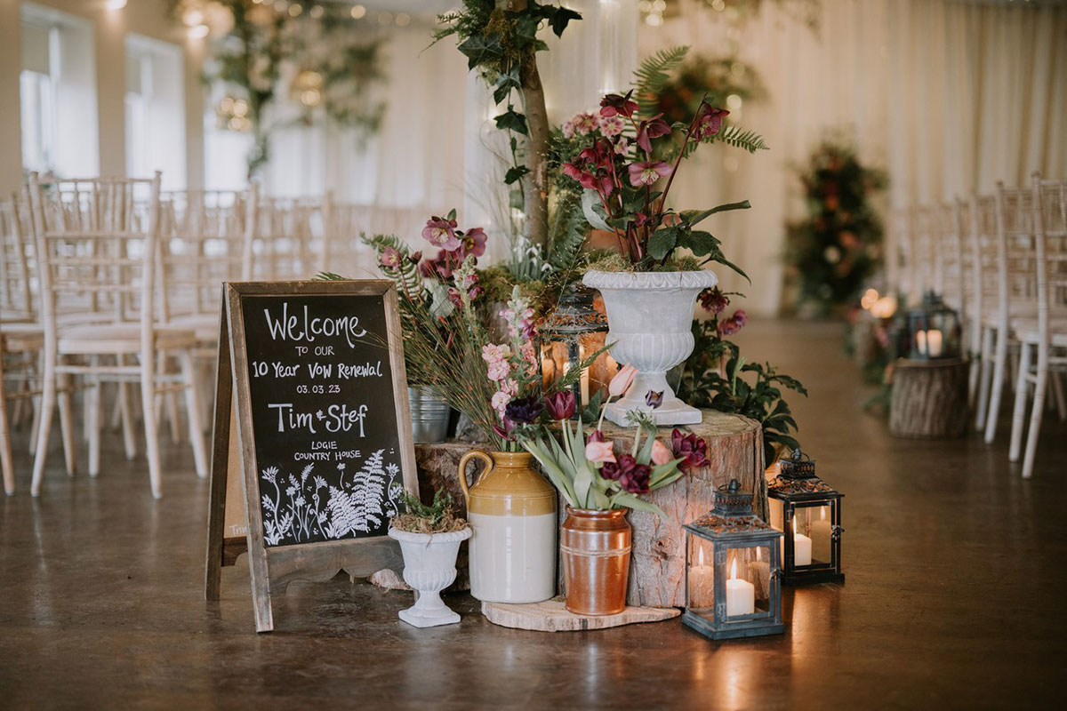 A ceremony entrance with a handwritten welcome sign, floral arrangements and lanterns beside rows of chiavari chairs.
