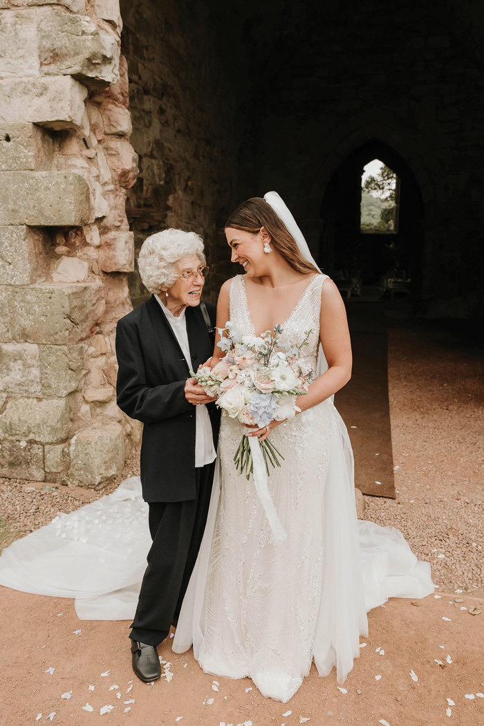 a bride posing for a photo with an elderly lady in front of a ruined stone building at Dunglass Estate