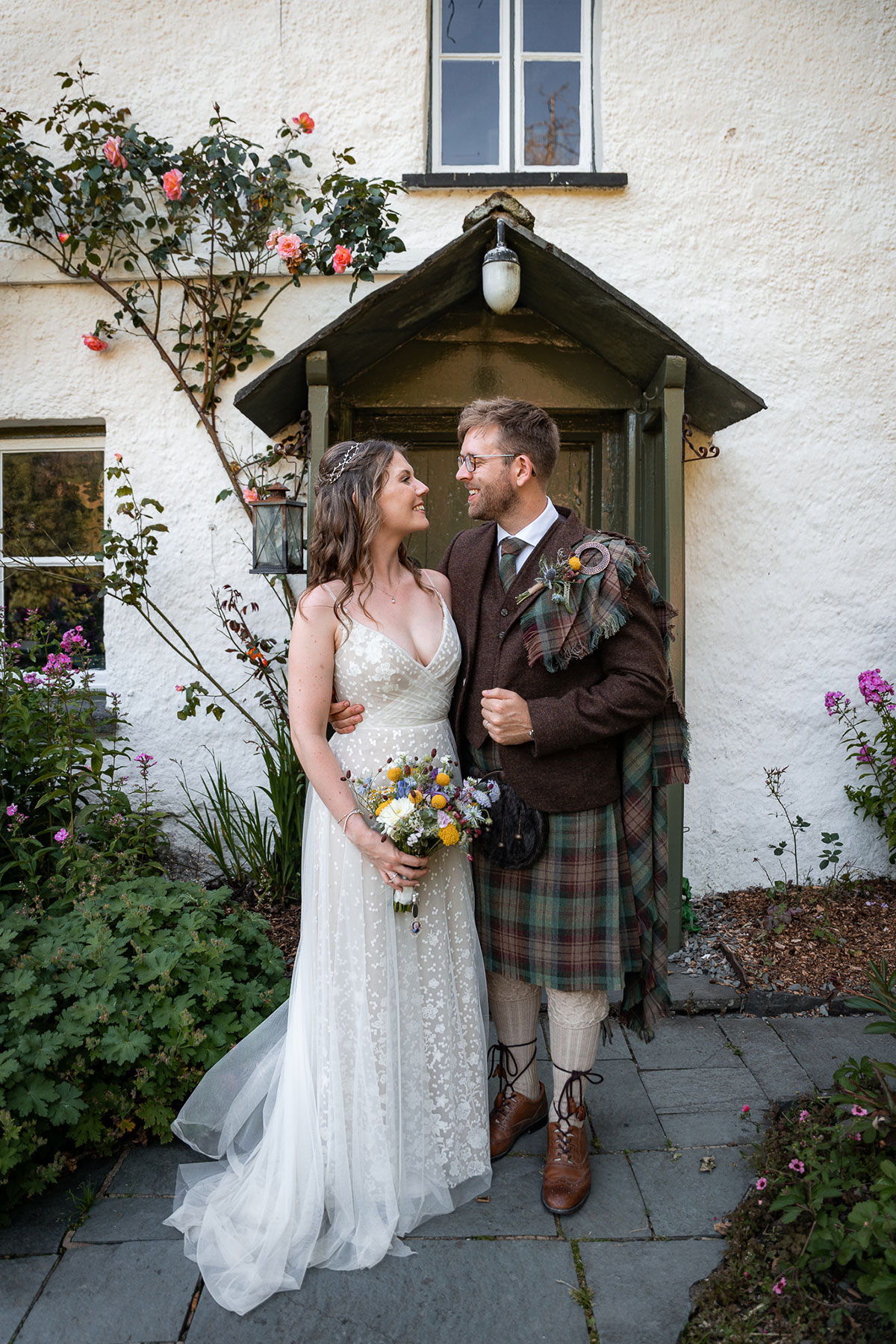 Bride and groom stand together outside a white cottage with climbing roses, smiling at each other. The bride wears a floral lace wedding dress and holds a colourful wildflower bouquet, while the groom wears a brown tweed jacket with tartan kilt and fly plaid