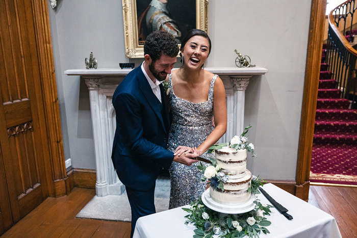 an elated bride wearing a silver glitter dress and groom wearing dark blue suit cut a two-tier semi-naked buttercream wedding cake that's sitting on a table in front of an elegant marble fireplace. There is a staircase with dark red carpet in the background