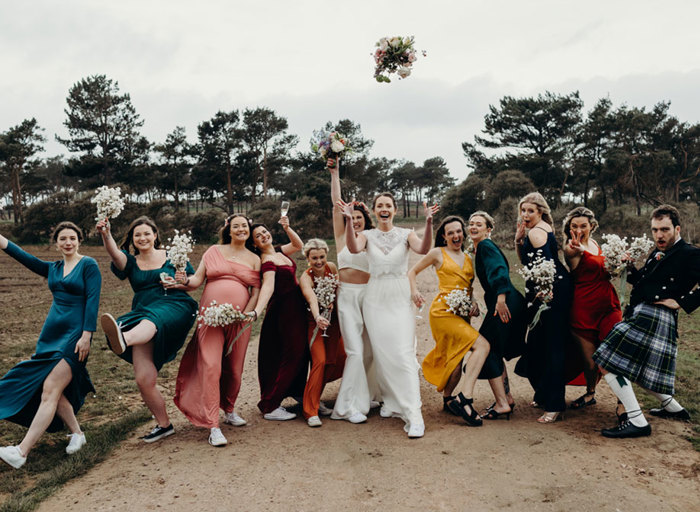 two brides and line-up of wedding guests wearing colourful attire posing for a photo on a sandy path with trees in background. One bride is throwing bouquet in the air and the second bride is holding her bouquet aloft