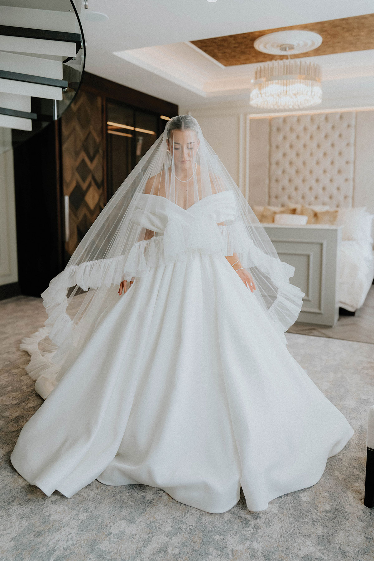 A bride wearing an off-the-shoulder ballgown and dramatic layered veil stands in a luxurious bedroom, softly lit by a chandelier