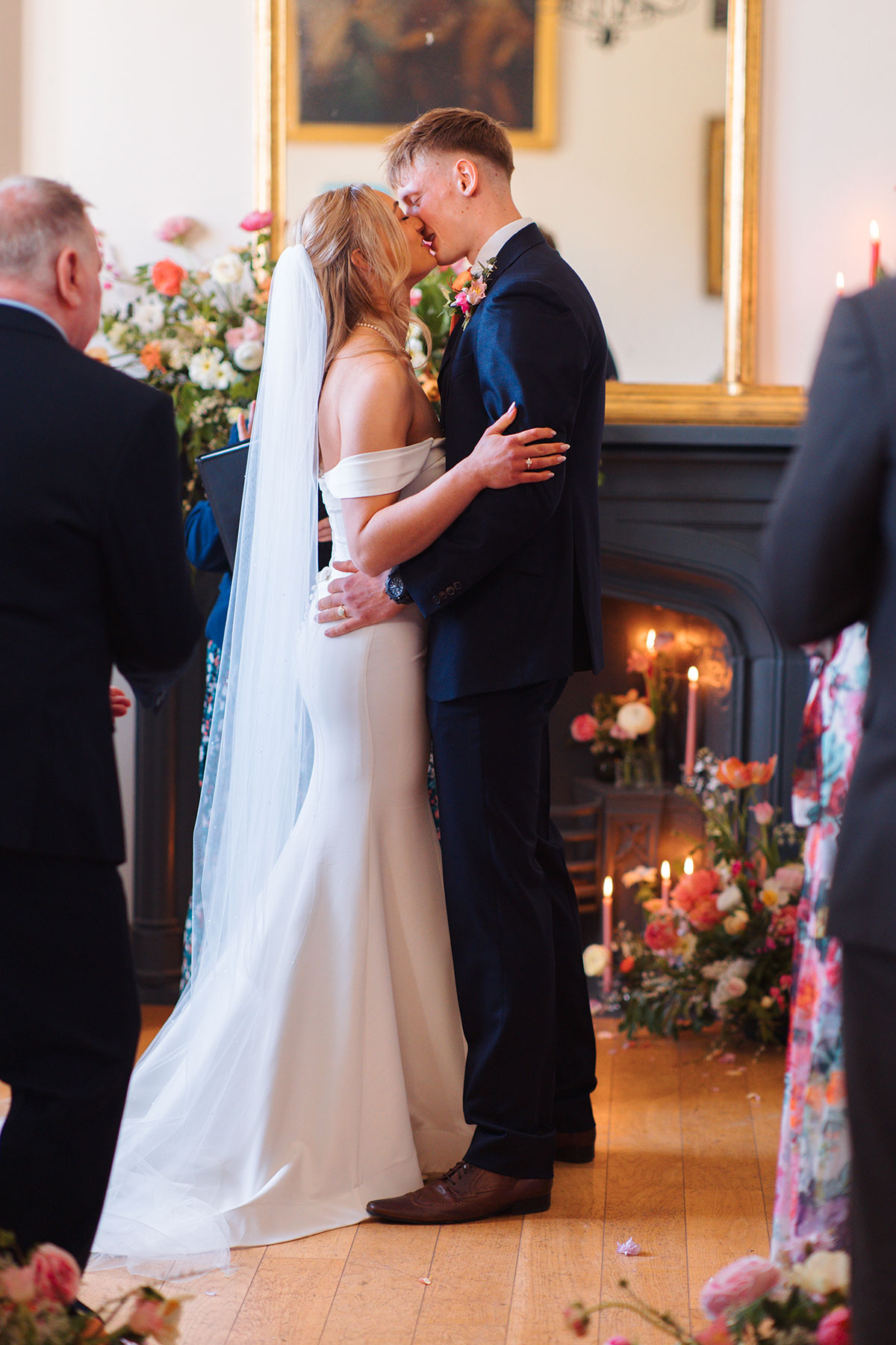 Bride and groom kissing during indoor ceremony with colourful floral backdrop and candles