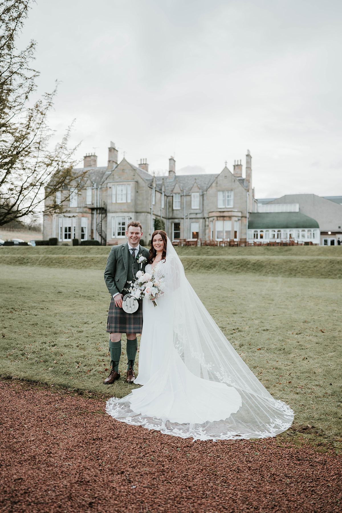 Couple posing outdoors in front of Norton House, bride holding bouquet with veil flowing on the grass