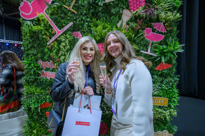 two people holding plastic champagne glasses standing in front of a fake grass wall that's covered in colourful cocktail decals