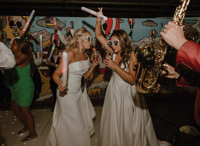 two brides wearing love heart sunglasses dancing with glow sticks on a light-up dance floor. They are both drinking cocktails through a straw. There is a saxophone in the foreground at the right hand side of the image
