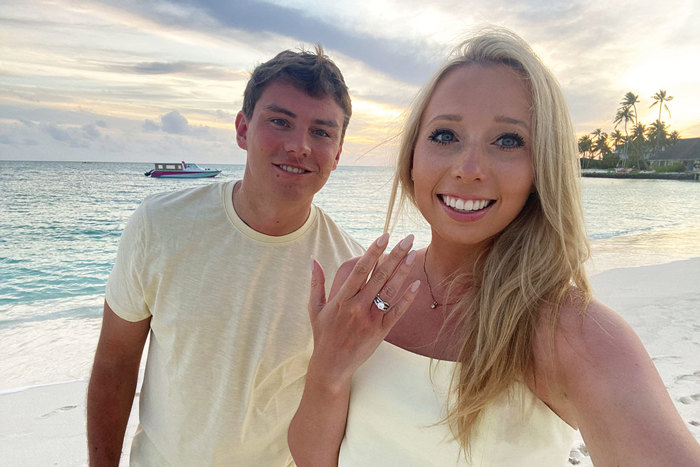 a couple on the beach in matching pastel yellow outfits, showing engagement ring to camera