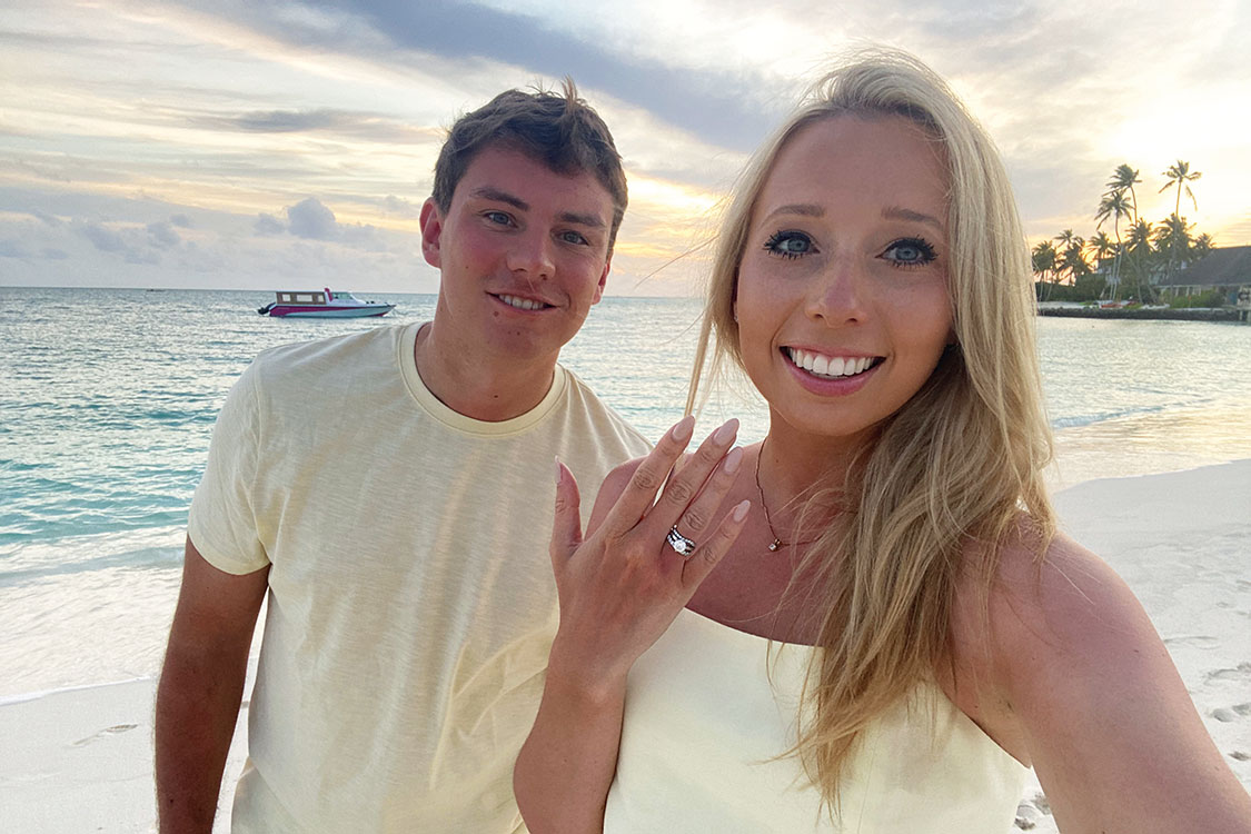 a couple on the beach in matching pastel yellow outfits, showing engagement ring to camera