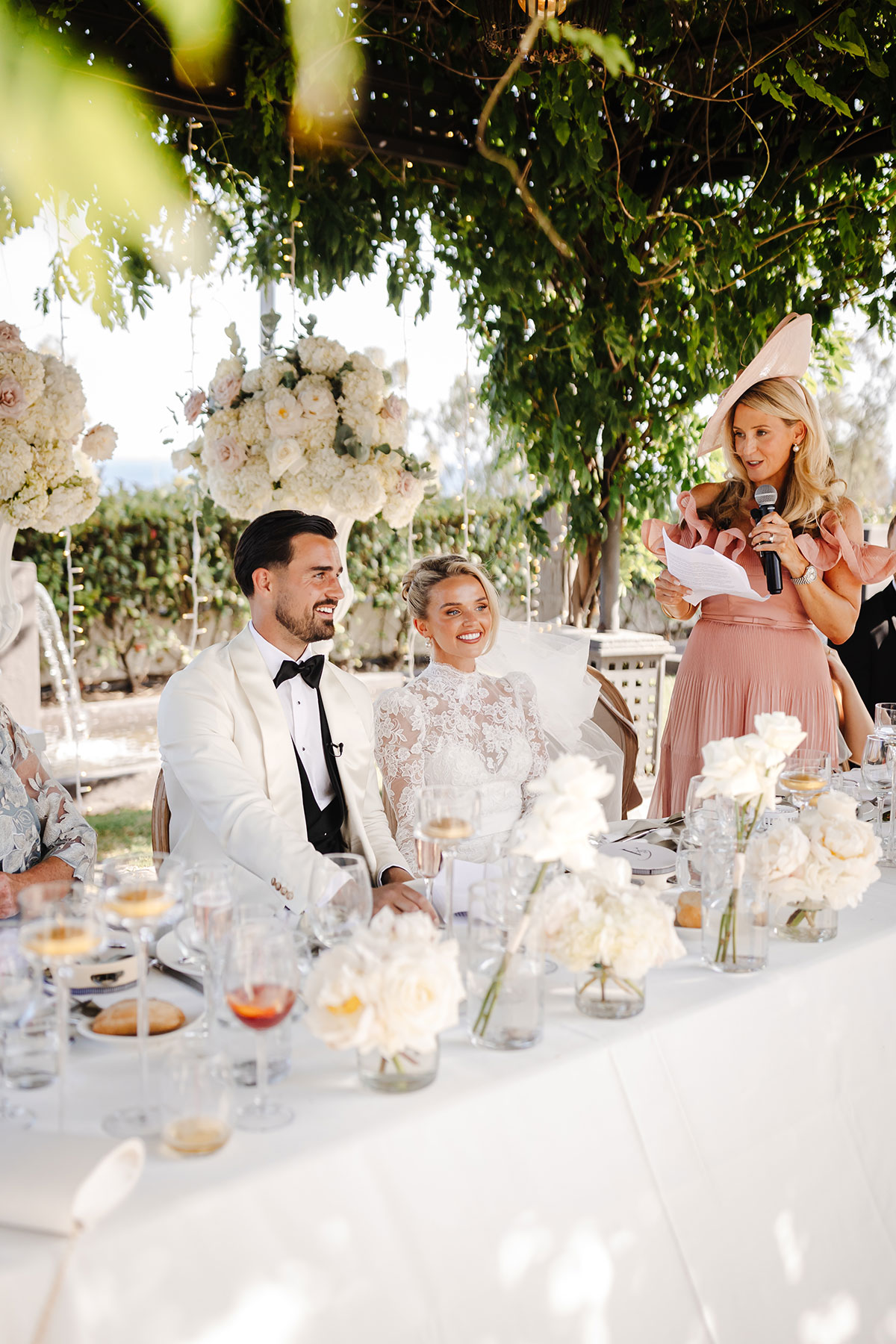 Bride and groom smile as a guest delivers speech at outdoor reception decorated with cream roses