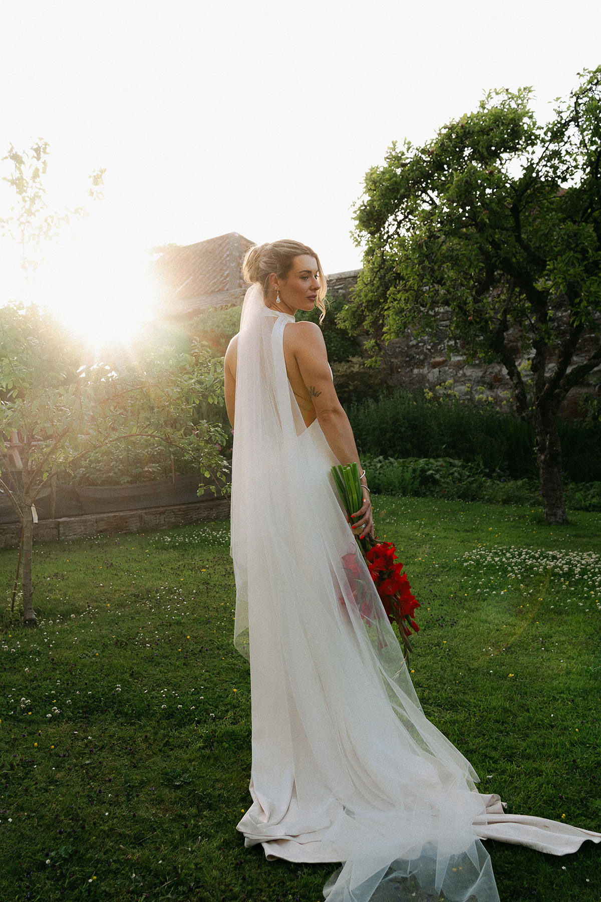 Bride stands in Kinkell Byre's orchard garden at sunset, wearing a minimalist wedding dress and long veil with a vibrant red bouquet.