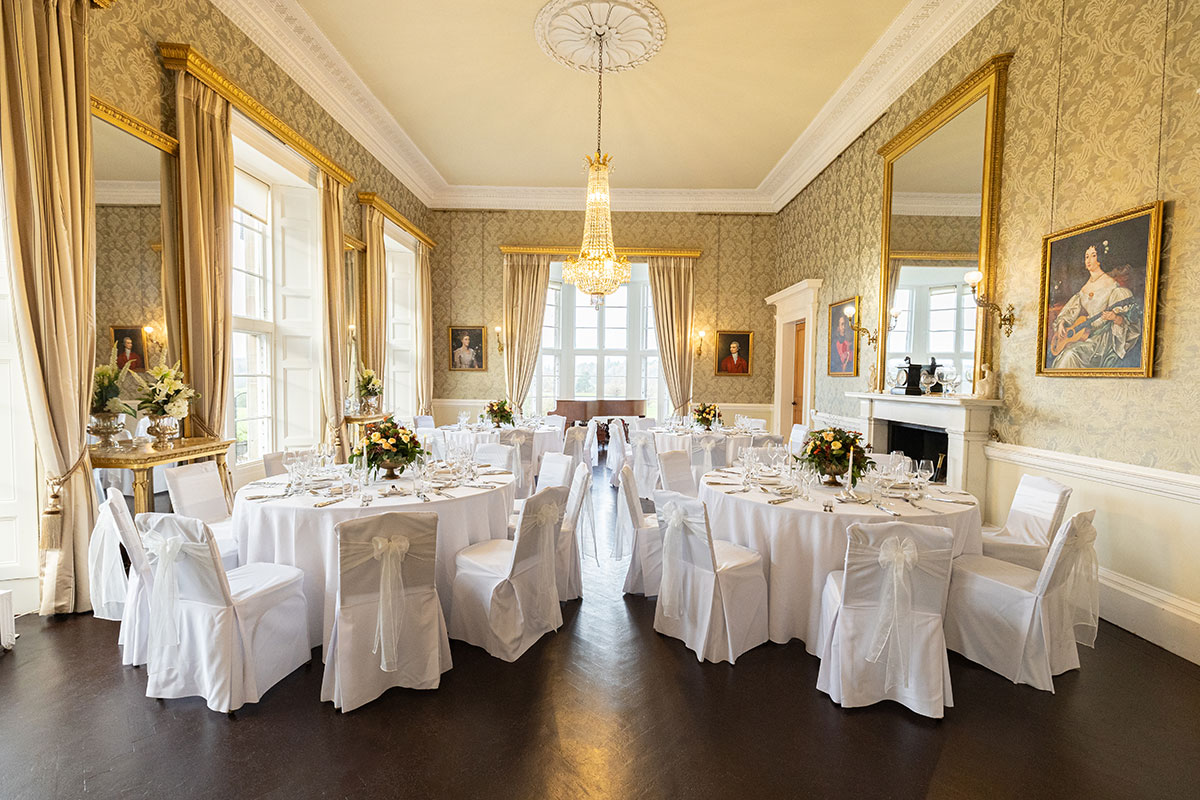 Formal wedding meal setup at Blairquhan Castle featuring round tables with white linen, floral centrepieces and chandeliers in a gold-toned dining room.