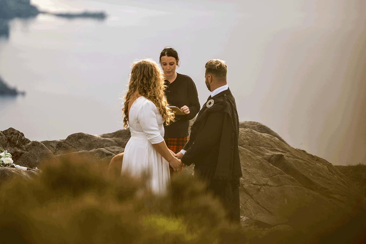 Couple hold hands during their outdoor wedding ceremony on a rocky mountain summit overlooking a Scottish loch at golden hour
