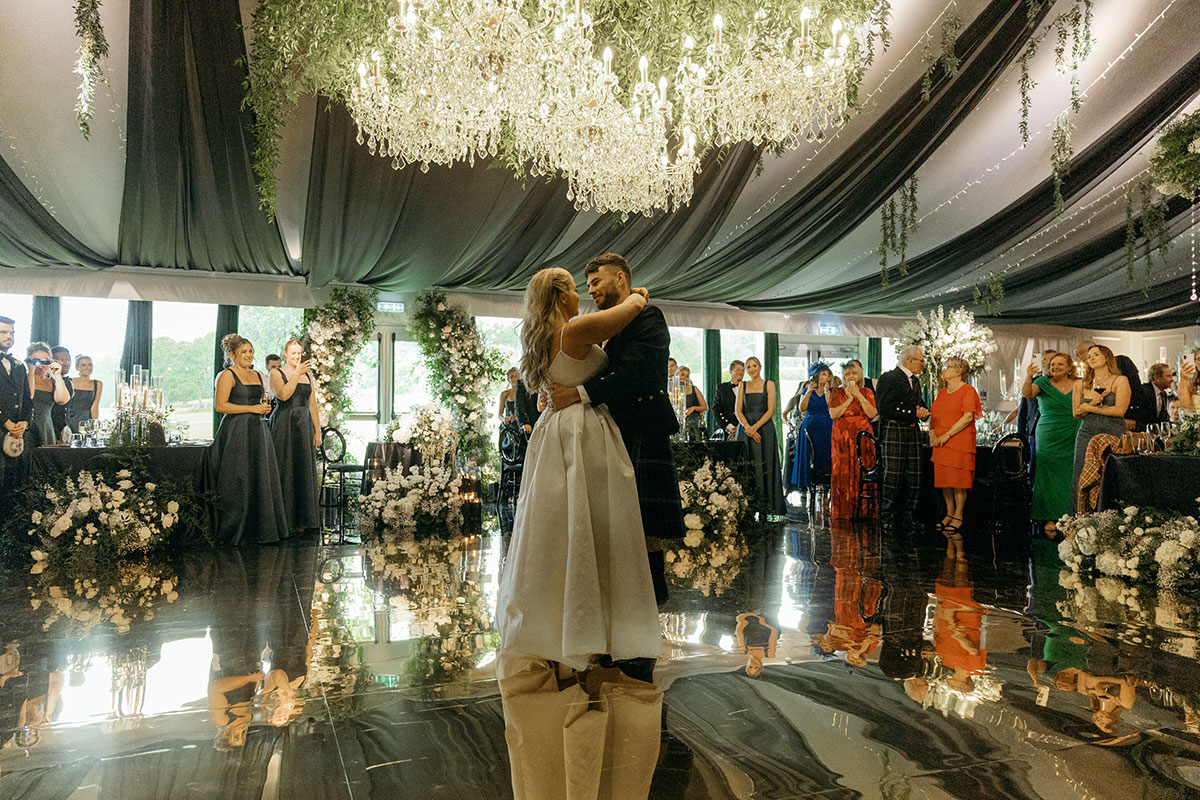 Couple’s first dance under chandeliers on mirrored floor surrounded by floral décor at Dundas Castle reception.