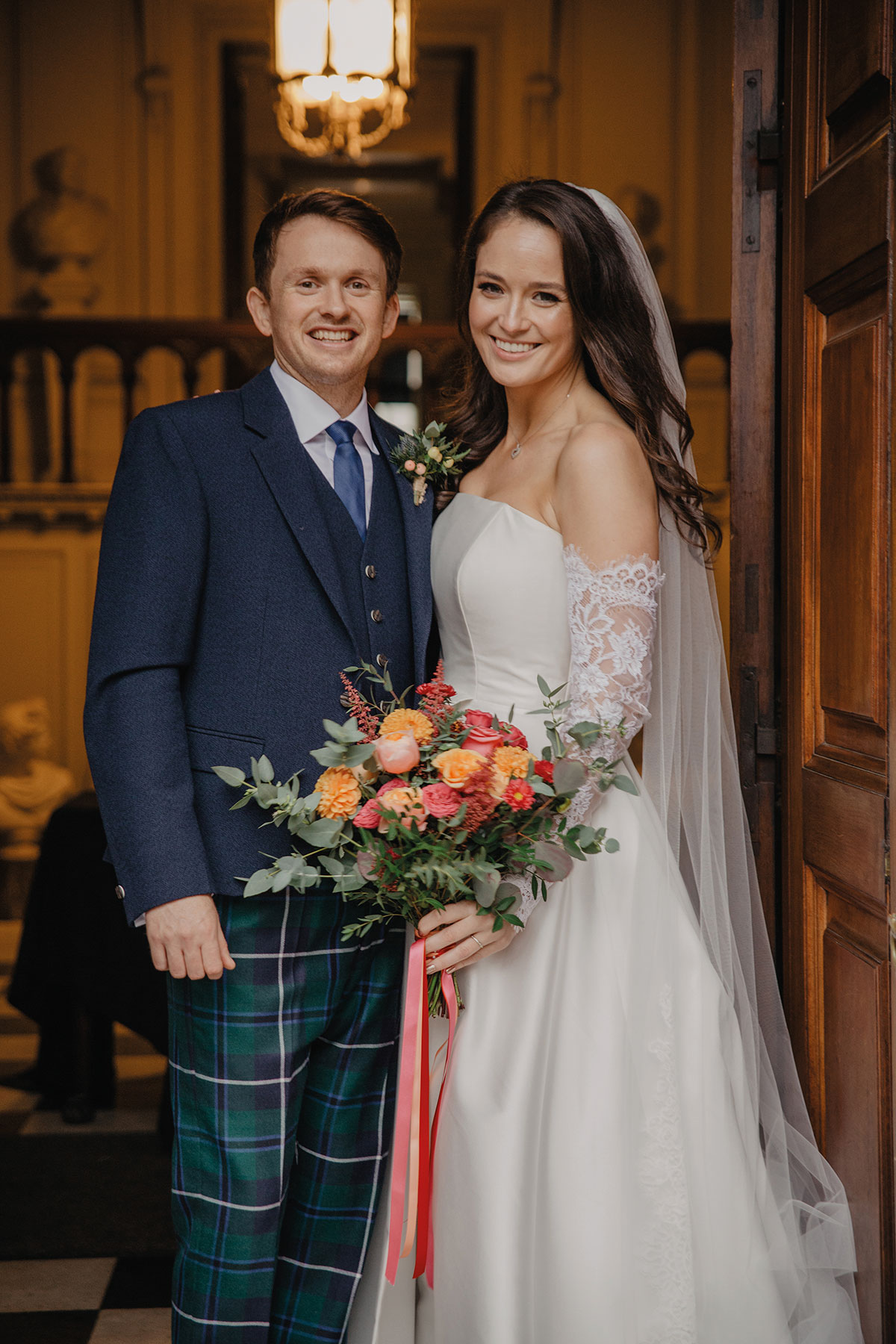 Bride and groom smiling in a doorway inside a historic venue; the bride wears an off-the-shoulder gown with lace sleeves and veil, and the groom wears a navy jacket with tartan trousers