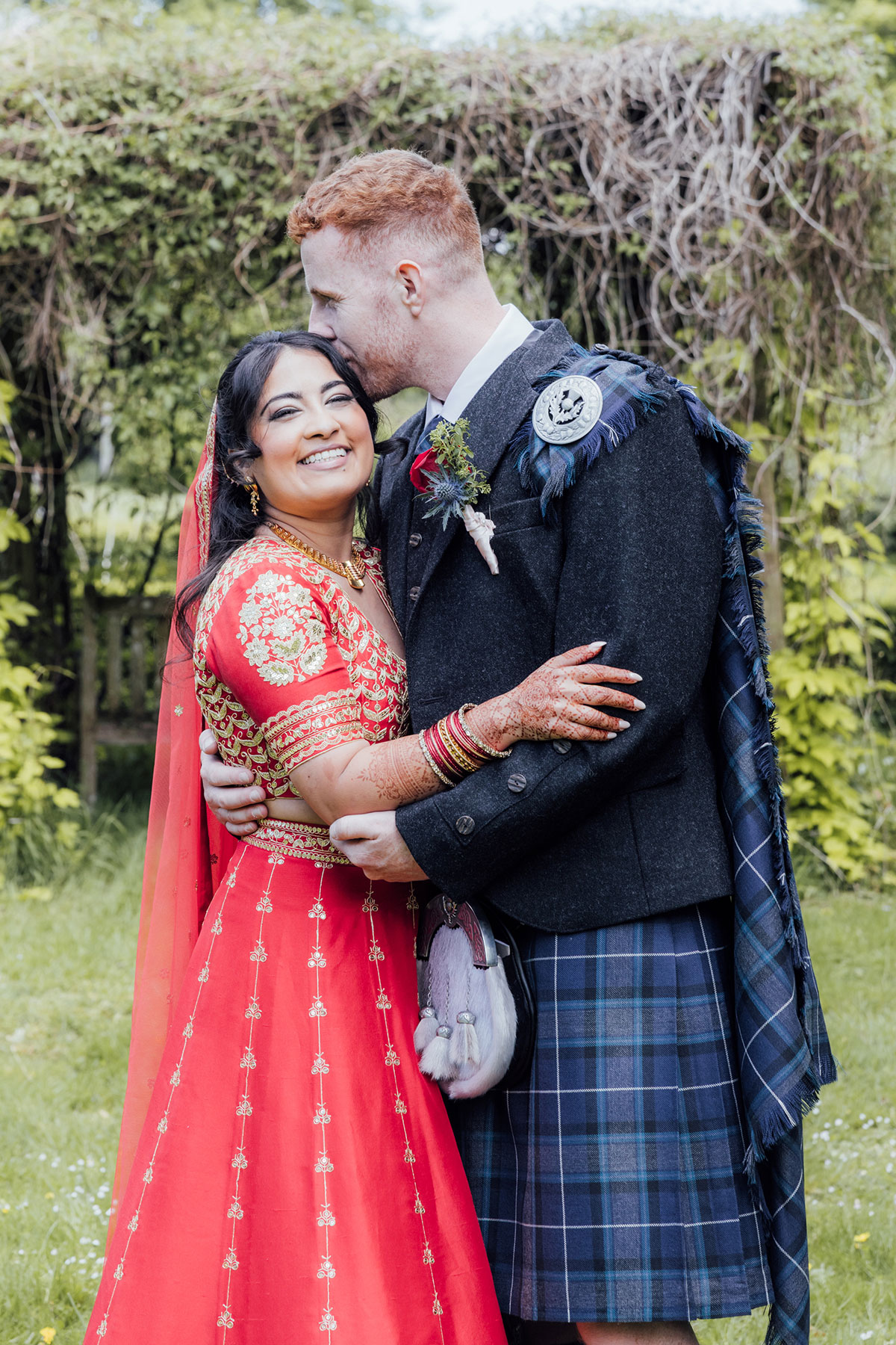Bride in a traditional red and gold Indian wedding dress embracing her groom in a Scottish kilt in the gardens of Atholl Palace Hotel