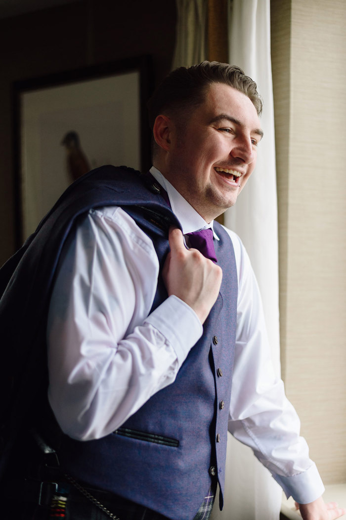 man in white shirt, blue waistcoat and purple tie, laughs as he tosses blue suit jacket over his one shoulder