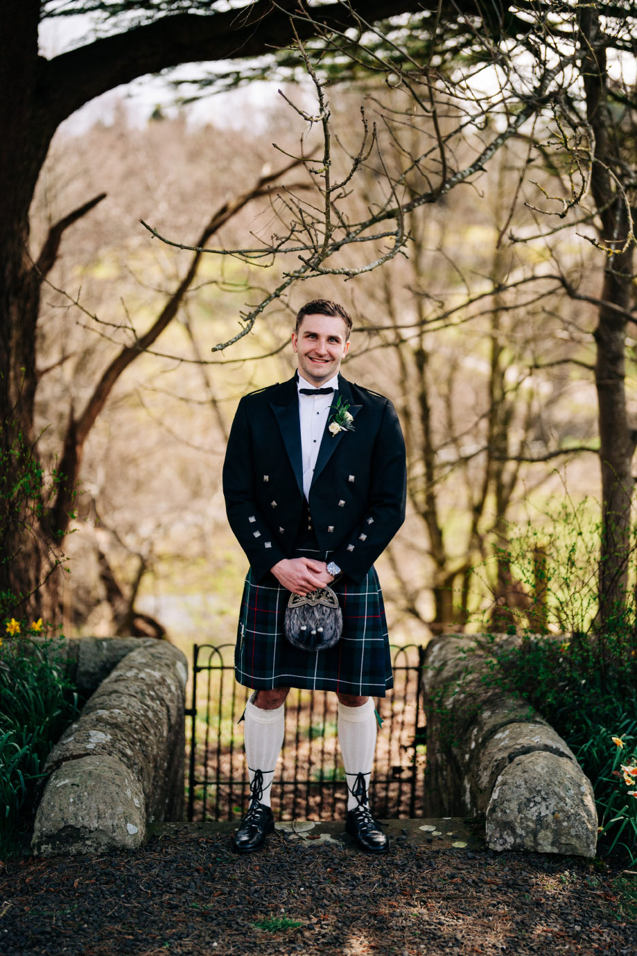 a man in a kilt standing below a wintry looking tree in front of a small wrought iron gate and small stone wall