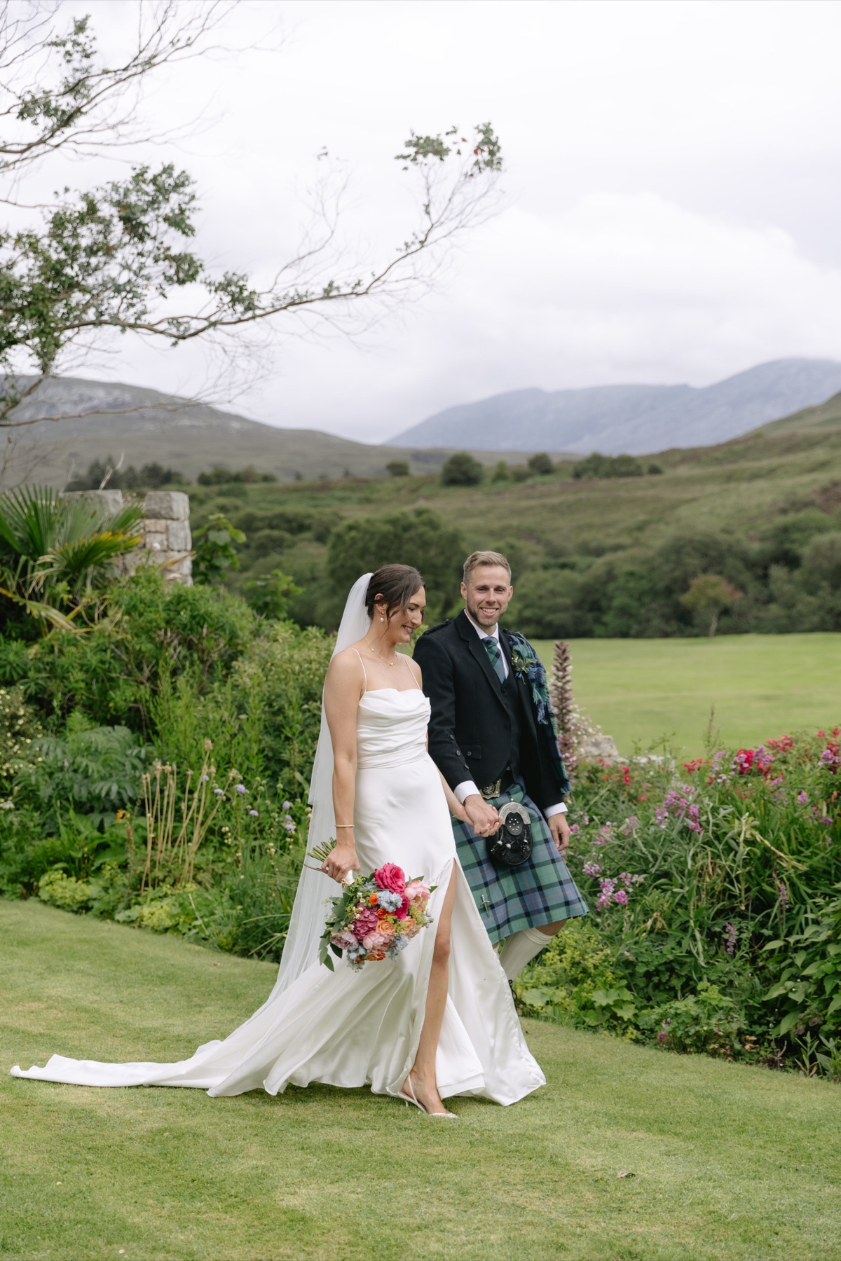 Newlyweds Gary and Isobel walk hand in hand through the gardens at The Dougarie Boathouse with Arran hills behind them.