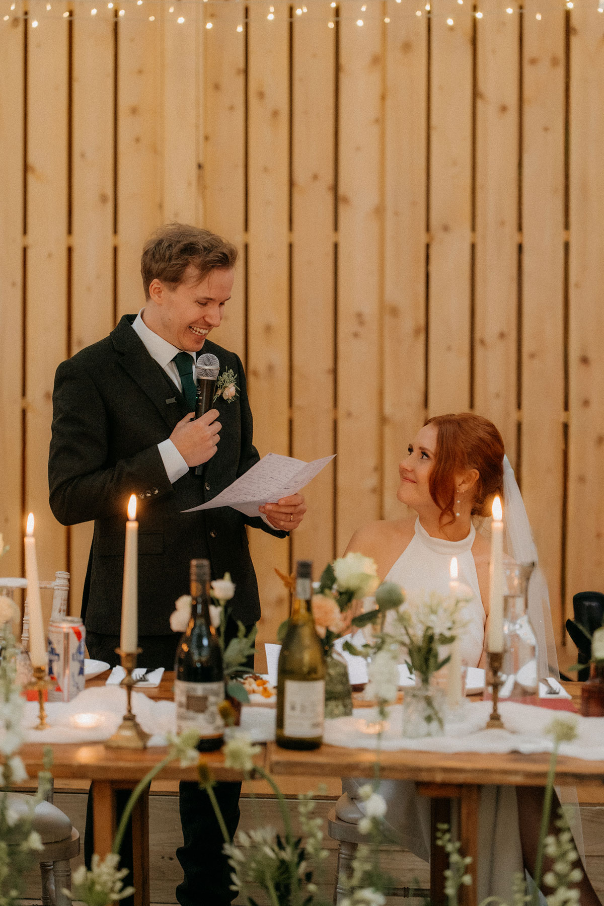 Groom reading wedding speech to bride at candlelit top table with floral centrepieces
