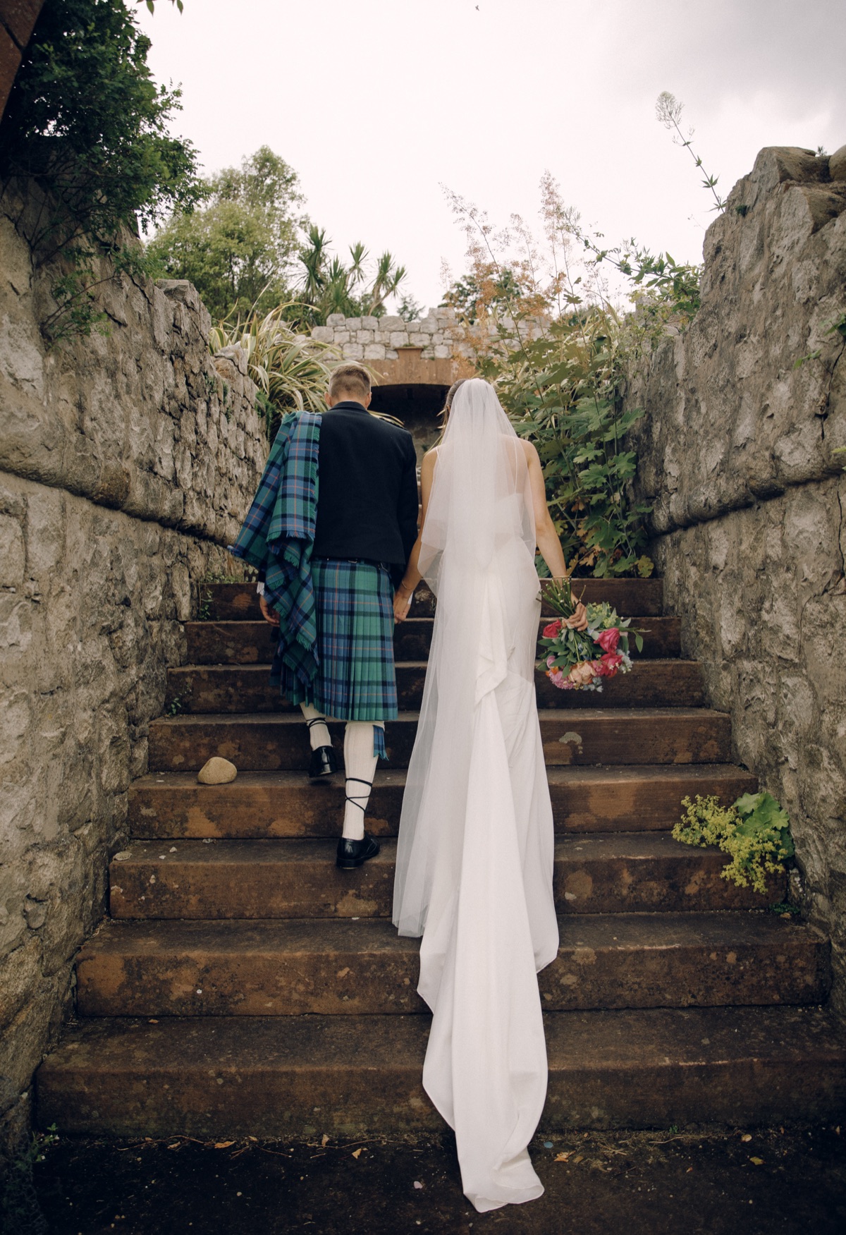 Newlyweds Isobel and Gary walk hand in hand up stone steps in the walled garden at The Dougarie Boathouse.