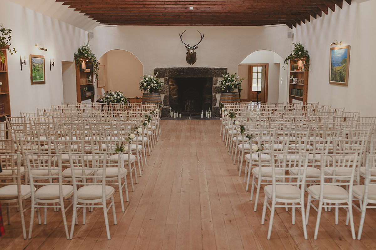 Rows of white chairs lining a wooden aisle in a rustic wedding ceremony room, with white and green floral arrangements surrounding a large stone fireplace
