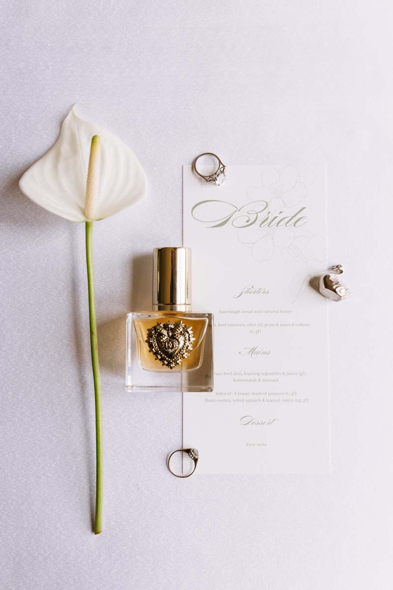 Cambo House wedding details featuring a white anthurium, bride’s menu, engagement and wedding rings, perfume and silver swan favour.