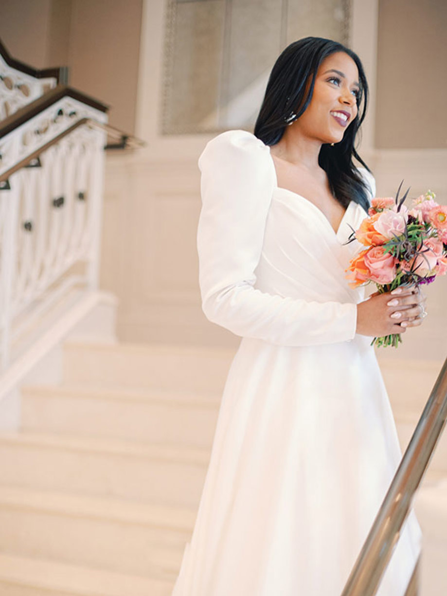 a bride in a long sleeved trailing gown stands on a staircase holding a pink and orange bouquet