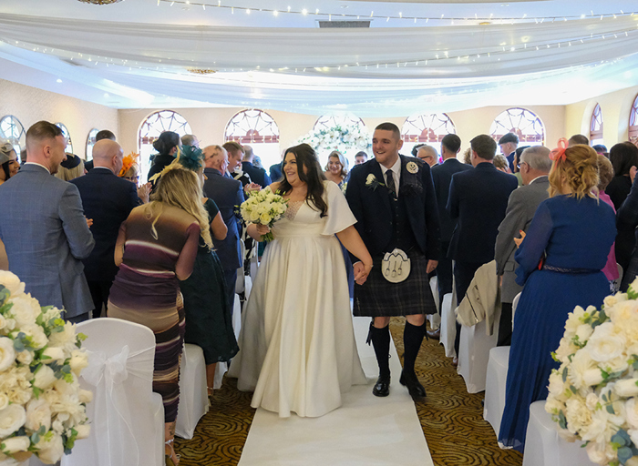 A bride and groom hold hands as they walk down the aisle together with their guests standing on either side of them