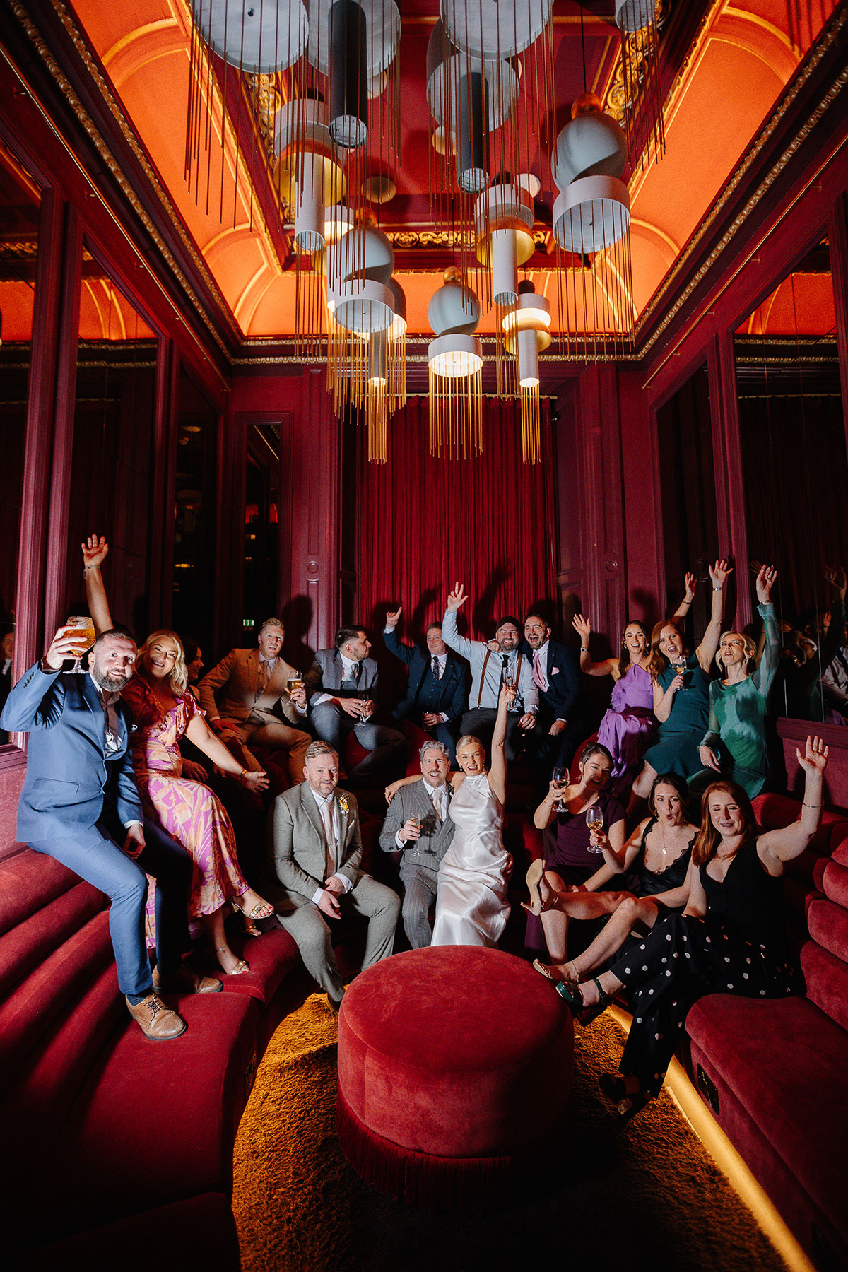 Newlyweds celebrate with champagne in the Scarlet Lounge at Virgin Hotels Edinburgh, sitting on red velvet seating surrounded by friends.